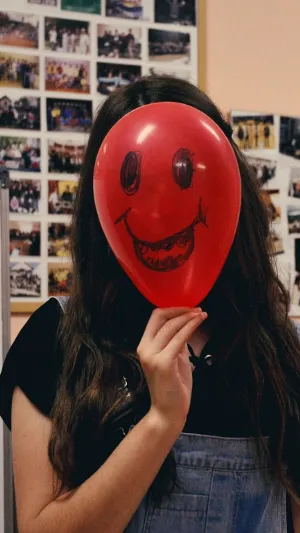 Image of a young women holding a baloon with a smiley face drawn on it in front of her own face.