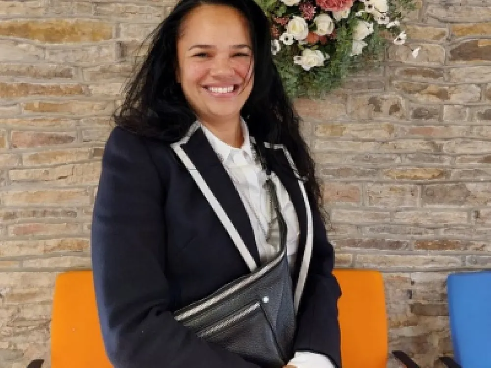 Photo of a woman against a stone wall with flowers. She is wearing a blazer and smiling.