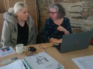 Photo of a young woman receiving advise from another woman who is sitting at a laptop.